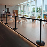 Retractable Queue Barriers for a modern, bright indoor cafe space with large floor-to-ceiling windows, light wood flooring, black stanchion barriers in the foreground, and several people seated at tables. 